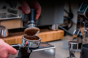 Tamper wird verwendet um den Kaffee runterzudrücken. Darunter ist die große Tamperstation zu sehen. Im Hintergrund ist eine Kaffeemaschine.